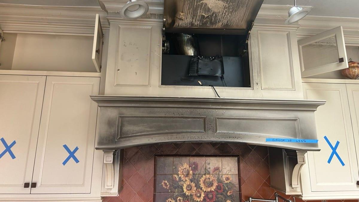 Soot-stained kitchen range hood marked as the cause of loss, with sampling locations taped on surrounding cabinets